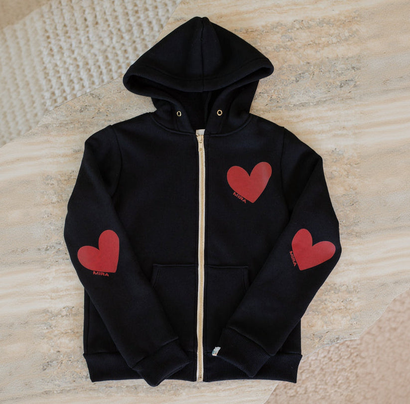 Black hoodie with red heart designs on sleeves and chest against a textured beige background