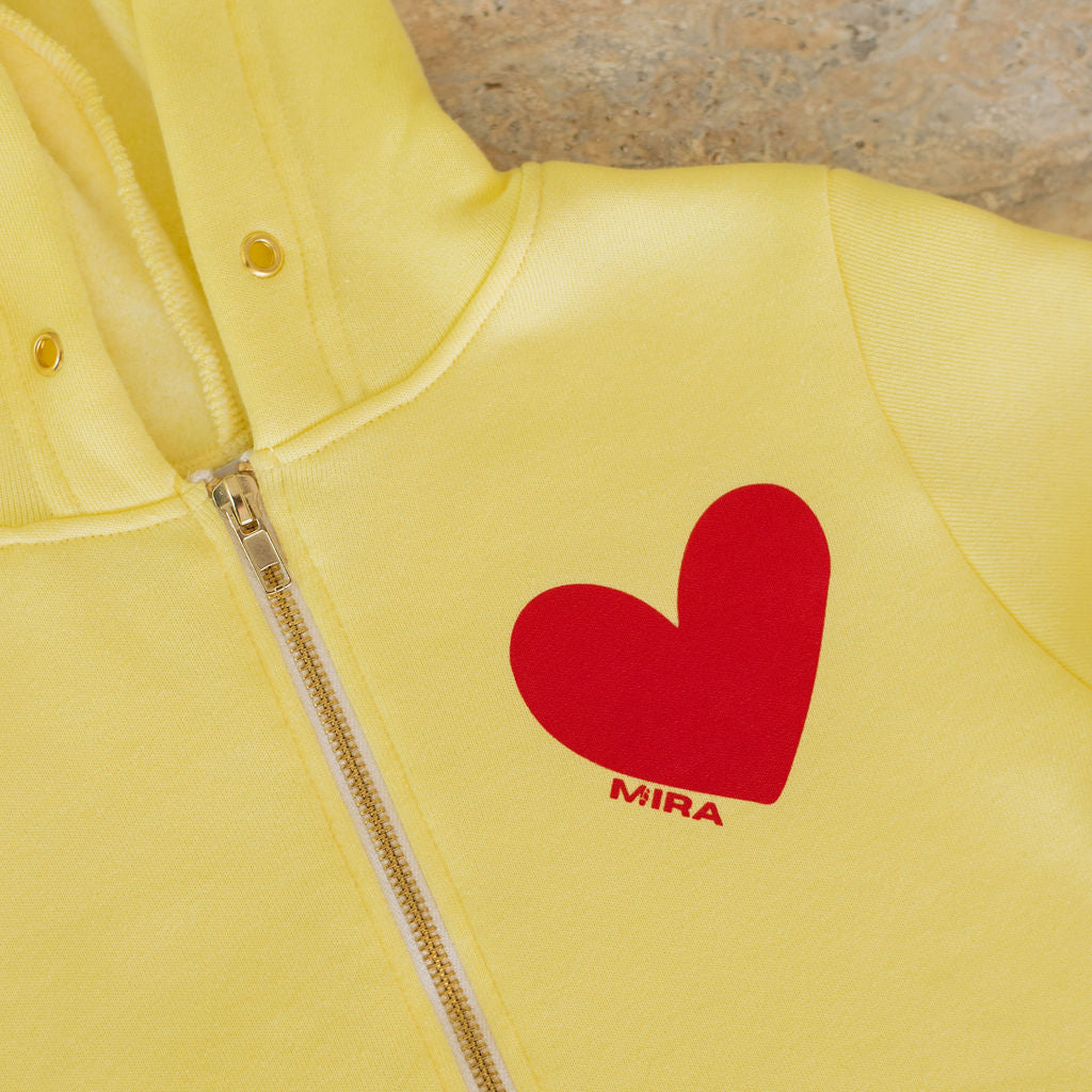 Yellow hoodie with a red heart and 'MIRA' logo on a stone surface