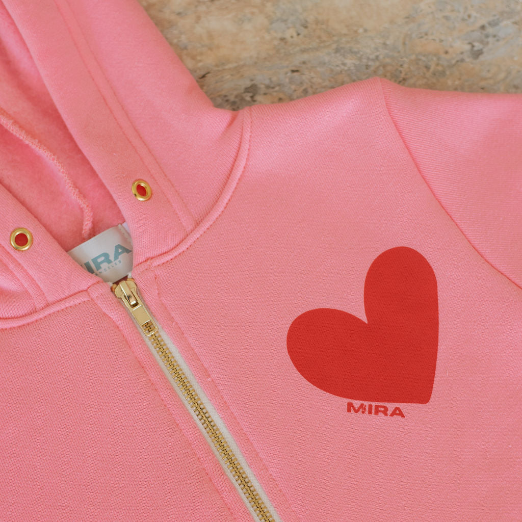 Pink hoodie with a red heart design and 'MIRA' branding on a textured surface.