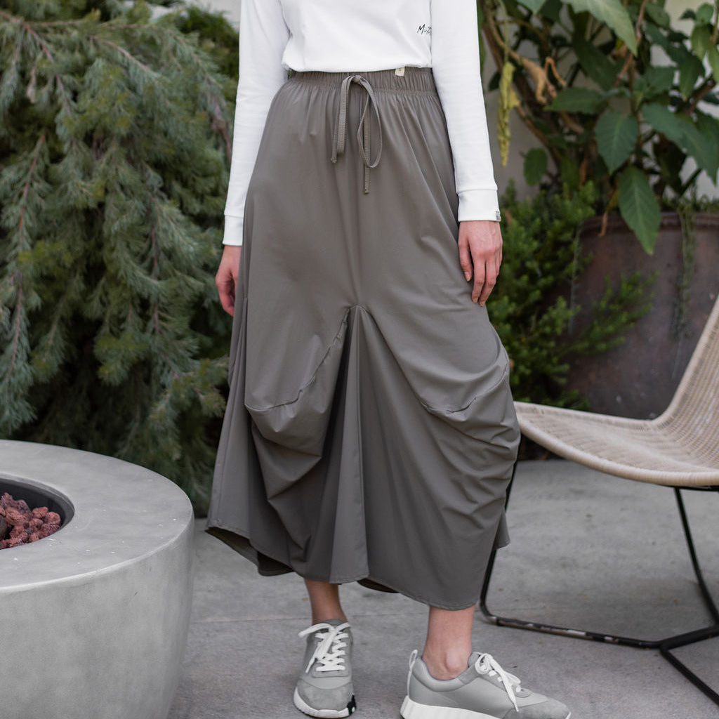 Woman wearing a white long-sleeve top and gray wide-leg pants standing outdoors with plants in the background.