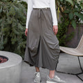 Woman wearing a white long-sleeve top and gray wide-leg pants standing outdoors with plants in the background.
