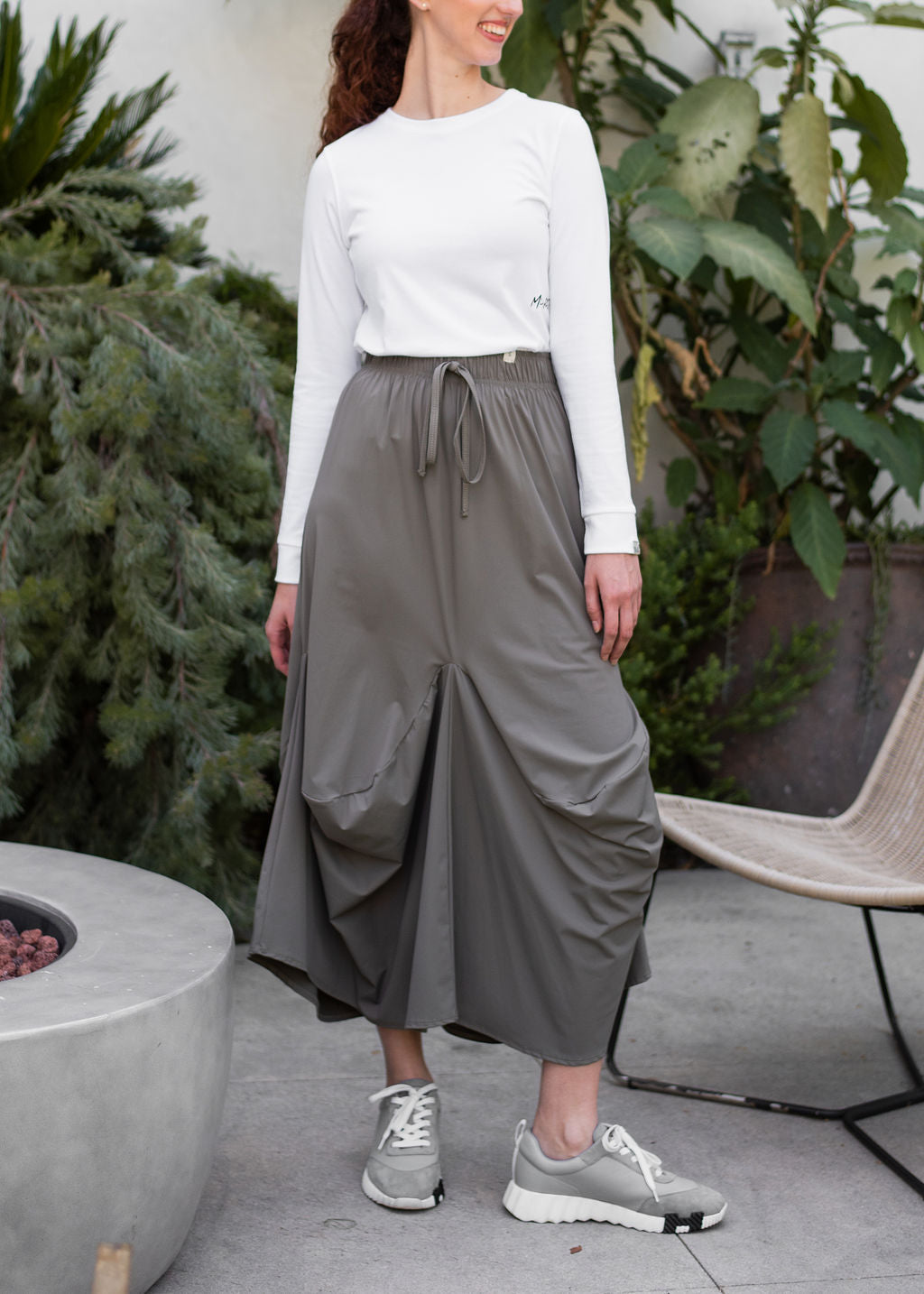 Woman wearing a white top and gray pants standing outdoors with plants in the background