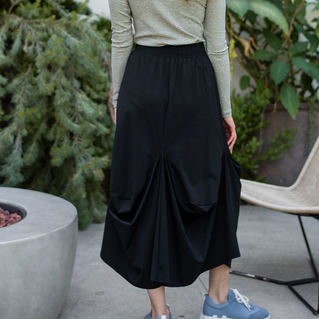 Person wearing a green long-sleeve top and black skirt standing outdoors with plants in the background.