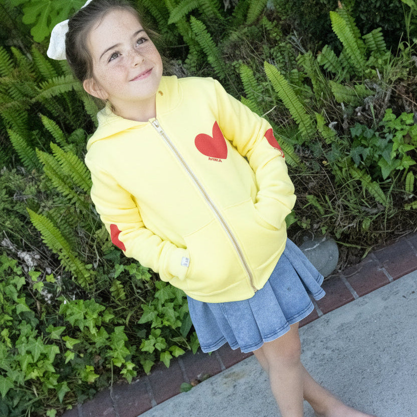 Young girl wearing a yellow jacket with red hearts, standing outdoors near greenery.